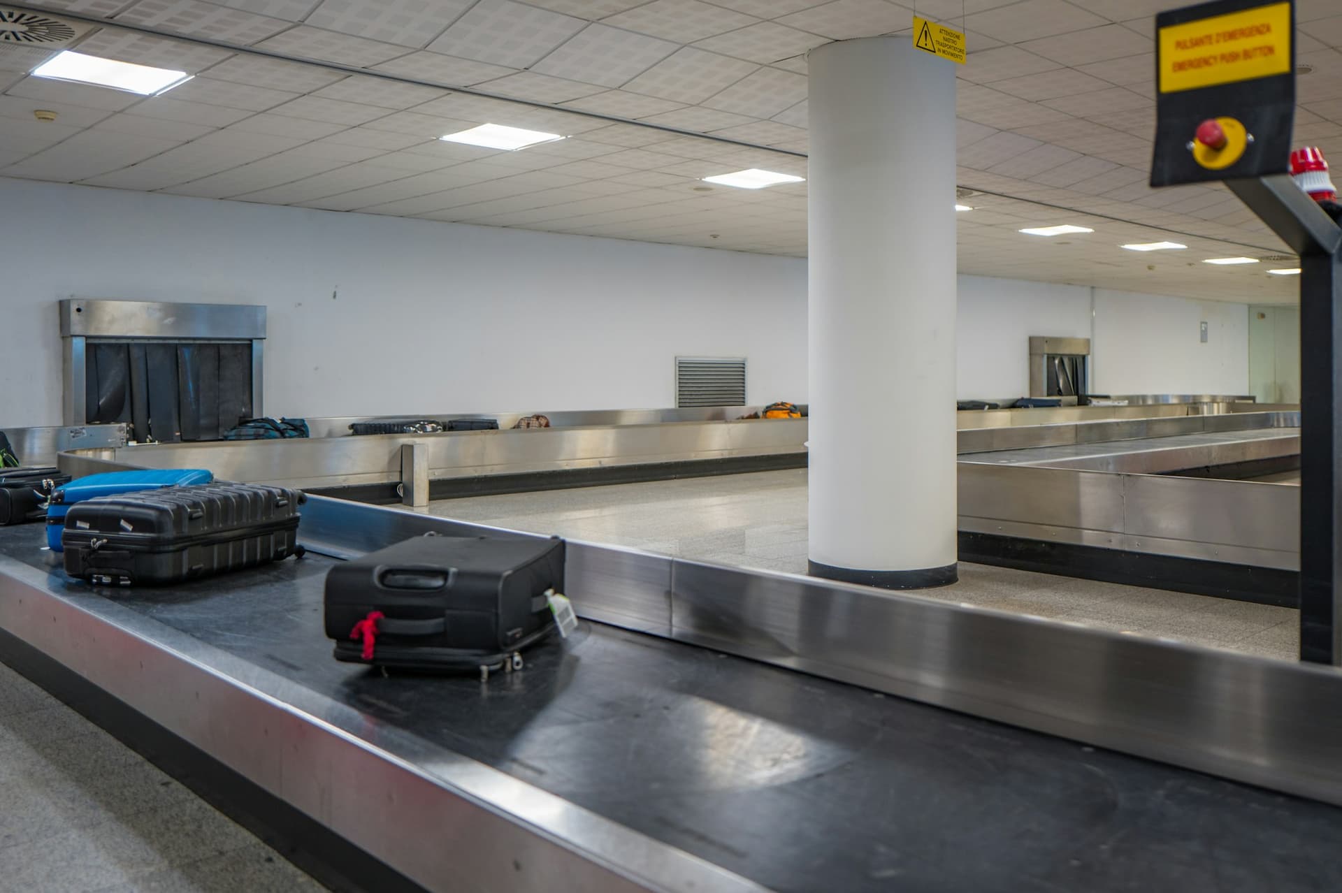 Un carrousel à bagages presque vide dans une zone d'arrivée d'aéroport. Trois valises tournent sur le tapis métallique ; un bouton d'arrêt d'urgence rouge est fixé au-dessus.